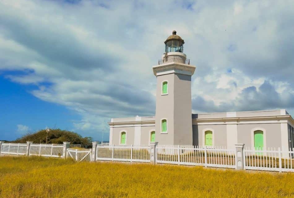 Faro gris con ventanas de color verde brillante rodeado de vallas blancas y césped amarillo.