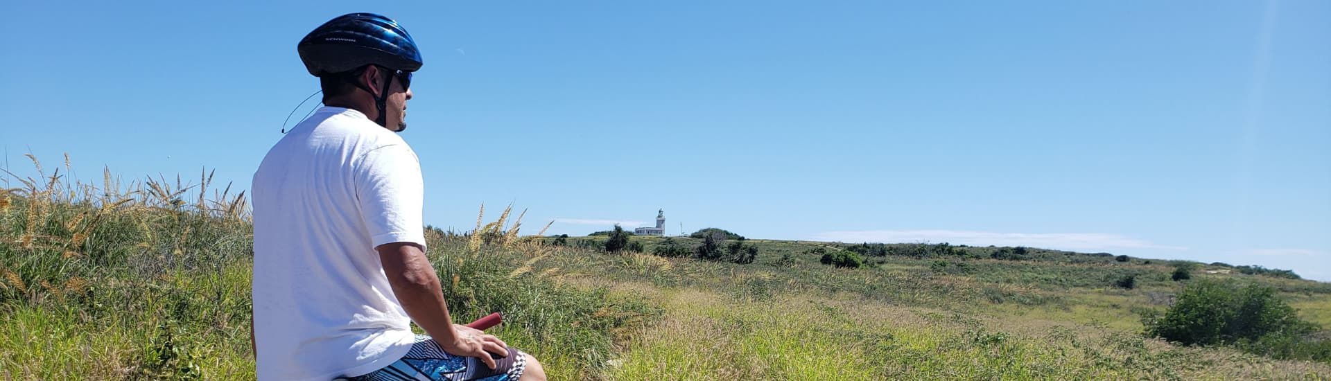 Man with helmet sitting on a bike overlooking a field of grasses and a lighthouse in the background