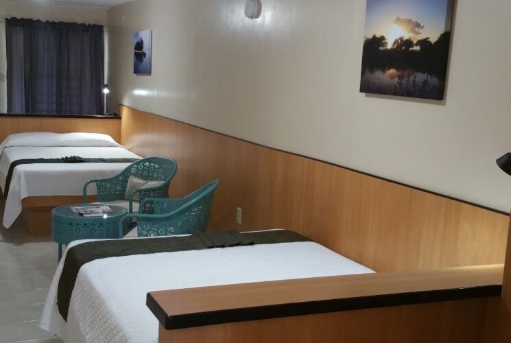 Bedroom with white walls with light brown wood paneling, tile flooring, two beds with white bedding, and sitting area