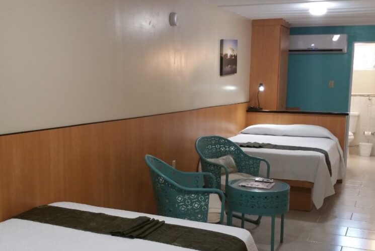 Bedroom with white walls with light brown wood paneling, tile flooring, two beds with white bedding, and sitting area