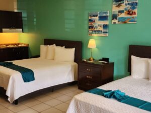 Studio with light green and tan walls, tile flooring, two queen beds with white bedding, dark wooden furniture, and full kitchen
