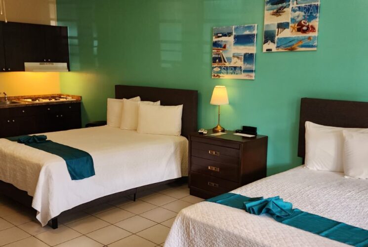 Studio with light green and tan walls, tile flooring, two queen beds with white bedding, dark wooden furniture, and full kitchen