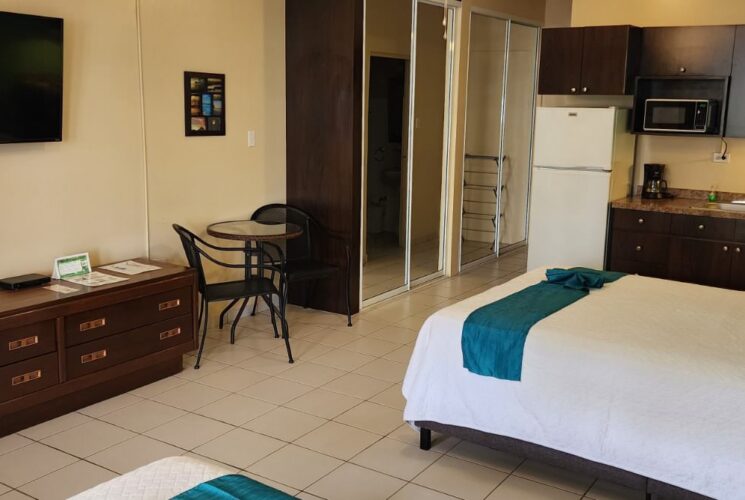 Studio with tan walls, tile flooring, two queen beds with white bedding, dark wooden furniture, flatscreen TV, and full kitchen