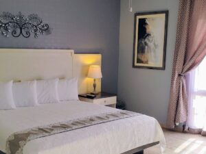Bedroom with light blue walls, tile flooring, king bed with white bedding, and double door with mauve and white curtains