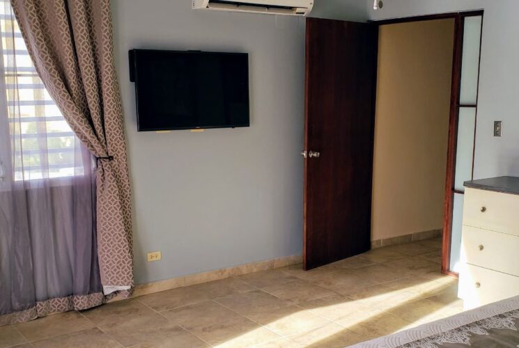 Bedroom with light blue walls, tile flooring, king bed with white bedding, white dresser, mirror, and flatscreen TV