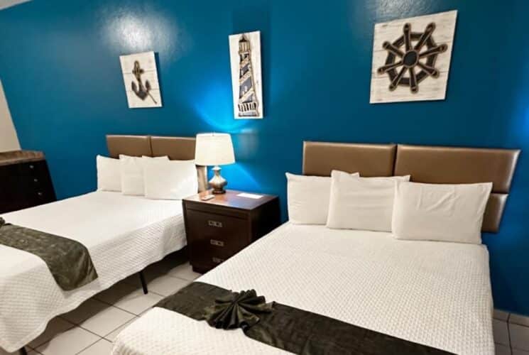 Bedroom with blue and tan walls, tile flooring, two queen beds with white bedding, and small counter with sink, microwave, and mini fridge
