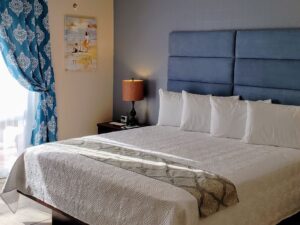 Bedroom with light blue and tan walls, tile flooring, blue upholstered headboard, white bedding, and large double door entrance with blue and white curtains