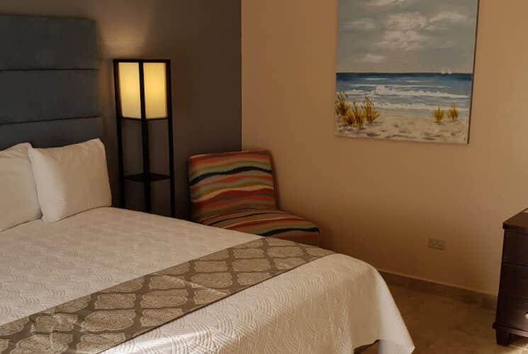 Bedroom with light blue and tan walls, tile flooring, blue upholstered headboard, white bedding, multicolored chair, and dark wooden dresser with mirror