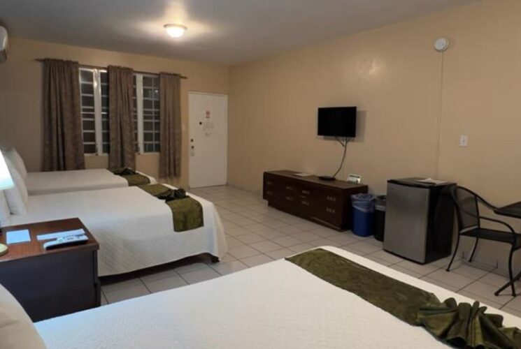 Bedroom with blue and tan walls, tile flooring, three beds with white bedding, flatscreen TV, mini fridge, and sitting area