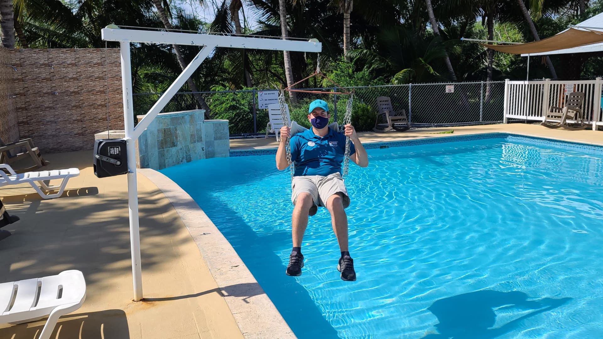 View of the pool surrounded by fencing and green vegetation with a person sitting in the handicap pool lift