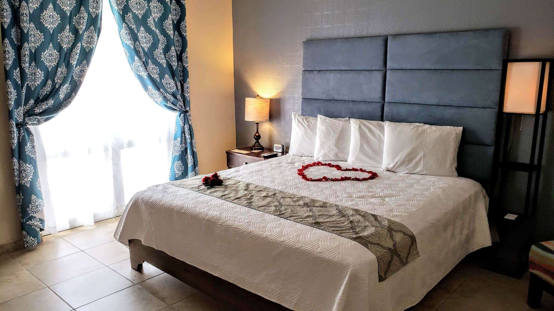 Bedroom with blue and cream walls, tile flooring, blue upholstered headboard, white bedding, and large double-door with white curtains and ornate blue curtains