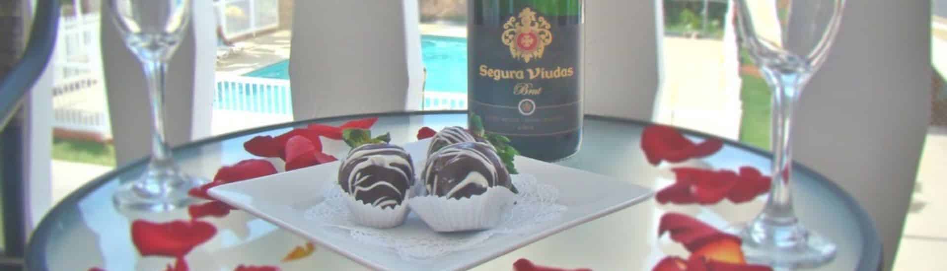 Vista de cerca de fresas cubiertas de chocolate en un plato blanco cuadrado, botella de champán, copas de champán y pétalos de rosa roja sobre una mesa de cristal