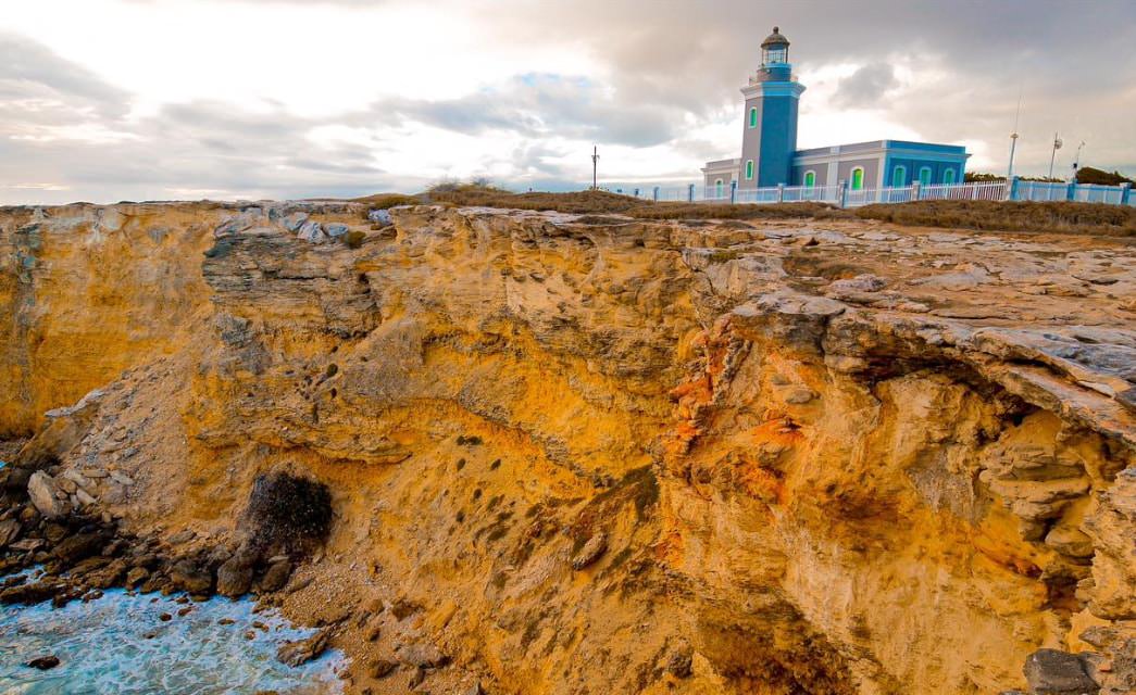 Faro gris con ventanas verdes brillantes rodeadas de vallas blancas en un alto acantilado