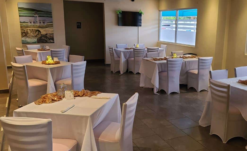Sala grande con paredes de colores claros, suelos de baldosas y varias mesas y sillas cubiertas con manteles y fundas blancas.