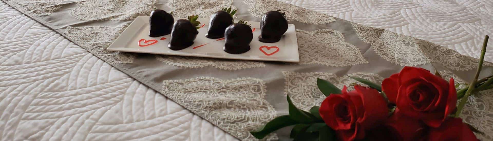 Close up view of chocolate covered strawberries on white rectangle plate and red roses on a bed with white bedding